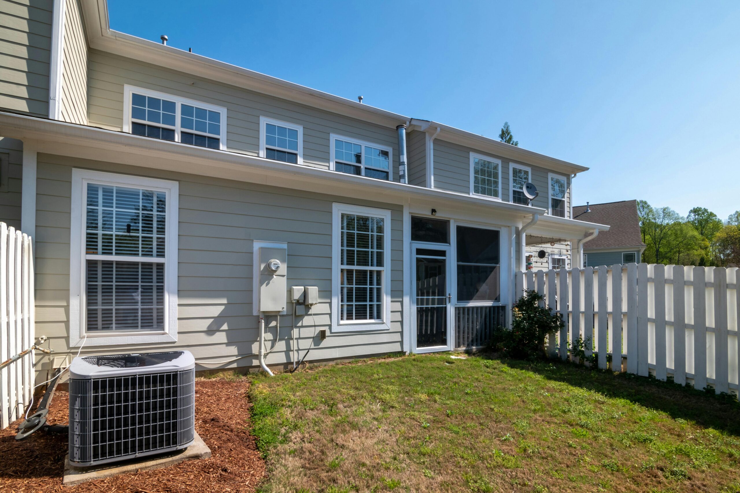 A sunlit backyard view of a modern grey house with white fencing and AC unit.